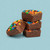 A stack of three square pieces of chocolate fudge topped with colorful M&M candies, with a fourth piece leaning against the base, set against a solid light blue background.