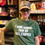 A smiling woman stands in a souvenir shop wearing a green t-shirt with bold white text that reads, "I WORK HARDER THAN AN UGLY STRIPPER." She is also wearing a camouflage hat with the phrase "MAKE URANUS GREAT AGAIN" printed on the front.