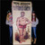 Two people holding up a large vintage style Tattooed Greek carnival sideshow banner from Uranus Fudge Factory