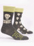 A pair of dark heather grey crew socks displayed against a plain white background. The socks feature a top cuff striped in lime green and cream. The front sock displays a large cream-colored cartoon skull and ribcage on the ankle. Knit into the side of the foot below it is bright lime green text that reads: "I ALMOST DIED but it WAS Just A COLD". The back sock shows that the rest of the design features an all-over pattern of smaller cream skulls.
