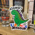 Green cartoon dinosaur sticker displayed on a wooden shelf beside a custom shot glass at Uranus Fudge Factory.