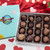 Blue gift box with sixteen assorted Uranus chocolates displayed on a table with hearts and roses in front of a heart background for Valentine's Day.