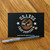 A black postcard on a wooden table featuring the 'Uranus Crop Dusters' logo — a vintage biplane flying over a brown circle, leaving a trail of tan cloud puffs behind it, flanked by two white stars. The website www.UranusFudgeFactory.com is printed at the bottom. A clear ballpoint pen lies in front of the card.