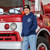 Man in navy Uranus Fire Dept hoodie with Maltese cross chest logo standing beside a vintage red Station 56 fire truck