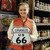 A smiling young woman in an orange quilted vest holding up a Uranus US 66 highway shield metal sign in a shop