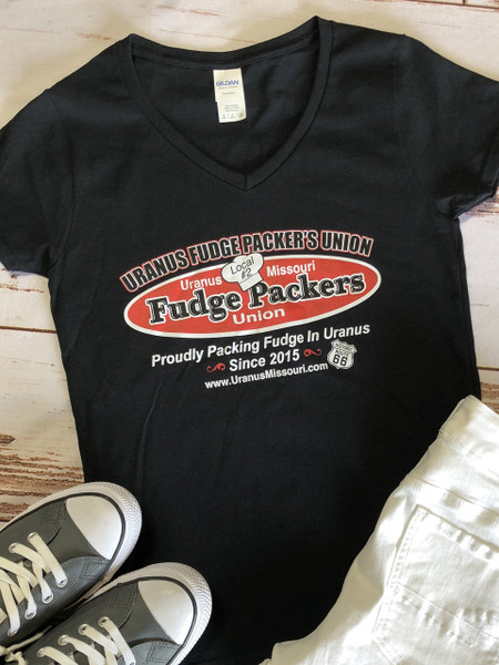 A flat-lay product photograph on a distressed light wooden background, centered on a black V-neck t-shirt. The chest graphic is clear, featuring the arching text: 'URANUS FUDGE PACKER'S UNION'. Below it, a red oval graphic contains the text 'Fudge Packers Union', with a chef's hat in the center marked 'Local #2'. Below the oval, text reads: 'Proudly Packing Fudge In Uranus Since 2015'. Next to this is a Route 66 shield icon.
