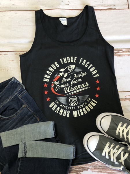 A flat lay photograph of a black tank top styled with cuffed dark denim jeans and gray canvas sneakers on a distressed white wood-plank background. The tank top features a retro, circular "Uranus Fudge Factory" logo in cream and red ink. The center displays a rocket ship orbiting the cursive slogan "The Best Fudge Comes from Uranus."