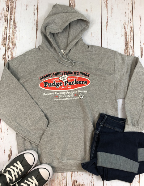 A flat lay of a heather gray pullover hoodie styled with folded dark denim jeans and gray canvas sneakers on a distressed white wood-plank background. The hoodie features the "Uranus Fudge Packer's Union" logo: a red oval containing the text "Fudge Packers Union" and a small white chef’s hat marked "Local #2."