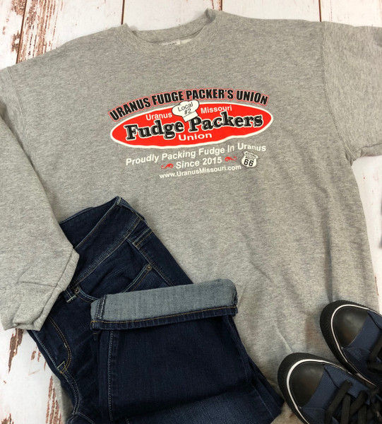 A flat lay of a heather gray crewneck sweatshirt styled with cuffed dark denim jeans and navy blue sneakers on a white wood-plank background. The sweatshirt features the "Uranus Fudge Packer's Union" logo, which includes a red oval with "Fudge Packers Union" in bold text and a small white chef’s hat marked "Local #2."
