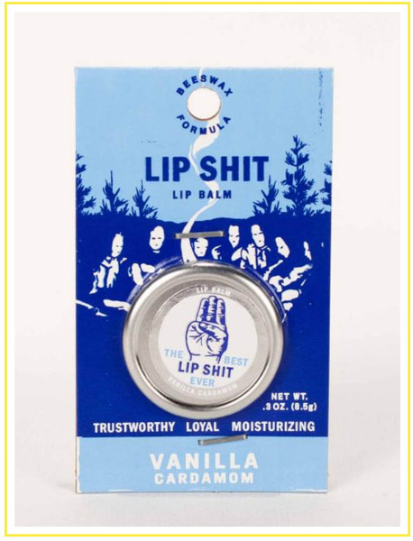 A retail package for "Lip Shit" lip balm in Vanilla Cardamom flavor. The blue and white card features a retro illustration of people in a forest, holding a circular silver tin that reads "The Best Lip Shit Ever" with a three-finger scout-style salute icon.