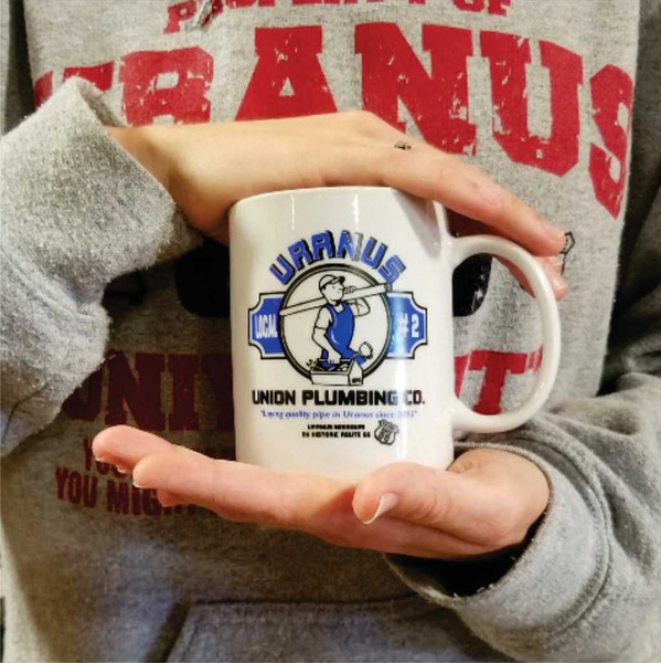 A person holding a white Uranus Union Plumbing Co. novelty ceramic mug from Uranus Fudge Factory.