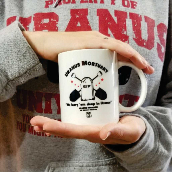 A person holding a white Uranus Mortuary novelty ceramic mug from Uranus Fudge Factory.