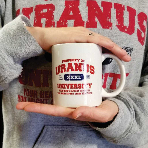 A person holding a white Uranus University ceramic mug from Uranus Fudge Factory.