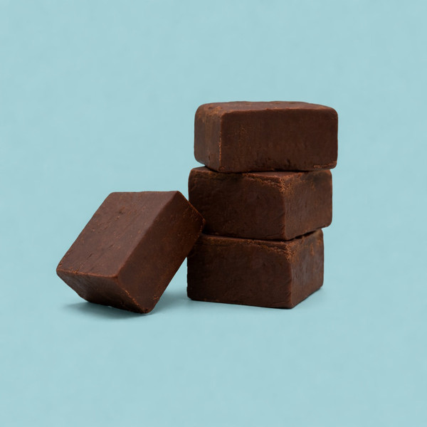 A stack of three thick squares of smooth, dark chocolate fudge, with a fourth square leaning against the base, is displayed against a solid light blue background.