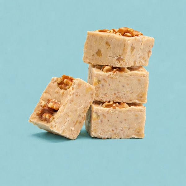 A stack of three square pieces of maple walnut fudge studded with chopped nuts, with a fourth piece leaning against the base, set against a solid light blue background.