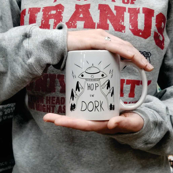 Person holding a white coffee mug with a black UFO graphic and a funny Hop In Dork quote.