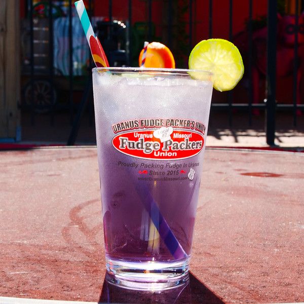 Purple drink in a Fudge Packers Union pint glass on an outdoor table with a fence background at Uranus Fudge Factory.