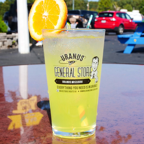 Yellow drink in a Uranus General Store pint glass on a table with a blurred parking lot background at Uranus Fudge Factory.