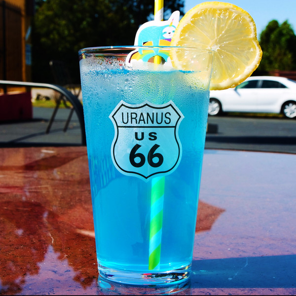 Blue drink in a Uranus Route 66 pint glass on a table with a blurred parking lot background at Uranus Fudge Factory.