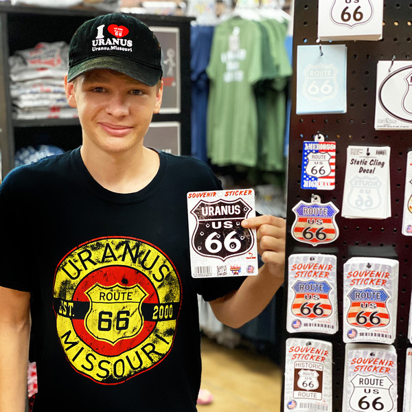 Smiling young man wearing a Uranus Missouri t-shirt and hat while holding a Route 66 souvenir sticker in a gift shop Uranus Fudge Factory.