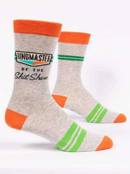 A pair of light heather grey crew socks displayed against a plain white background. The socks feature bright orange accents on the toe, heel, and top cuff, along with two thin green stripes on the upper calf and three thicker lime green stripes across the foot. Knit into the ankle area of the front sock is a retro-style graphic with the word "RINGMASTER" in tall black letters inside an outlined box, underlined by geometric teal and orange shapes. Below the graphic, black text reads "OF THE" in small caps, followed by "Shit Show" in a bold cursive script.