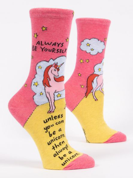A pair of crew socks displayed against a plain white background. The upper half of the socks is bright pink, while the lower foot section is yellow, with matching pink accents on the toe and heel. The design features a pink unicorn with a red mane standing in front of a fluffy light blue cloud, surrounded by small yellow stars.