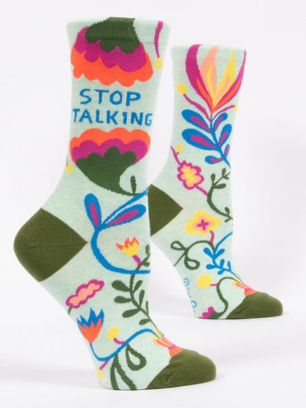 A pair of light mint green crew socks displayed against a plain white background. The socks feature olive green accents on the toe, heel, and top edge. They are decorated with a vibrant, bold floral pattern featuring stylized vines, leaves, and large flowers in bright pink, orange, blue, and yellow. Knit into the upper ankle of the front sock, sandwiched between two large colorful floral shapes, is the blue block text: "STOP TALKING".