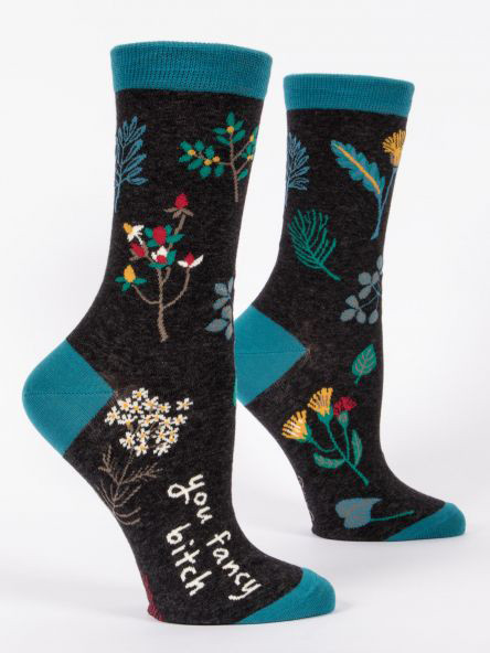 A pair of dark charcoal grey crew socks displayed against a plain white background. The socks feature teal accents on the toe, heel, and top cuff. They are decorated with a scattered botanical pattern of various green leaves and delicate red, yellow, and white flowers. Knit into the side of the foot on the front sock is the white script text: "You fancy bitch".