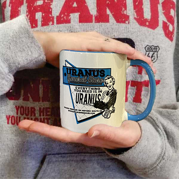 Person holding a cream Uranus General Store coffee mug with a blue handle from Uranus Fudge Factory.