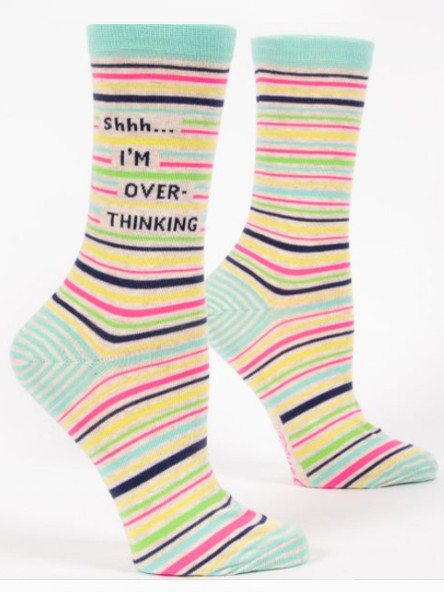 A pair of cream-colored crew socks displayed against a plain white background. The socks feature a solid light mint green top cuff and a heel section striped in mint green and white. The main body of the sock is covered in a pattern of horizontal stripes in varying widths, featuring bright pink, yellow, lime green, mint, and dark navy blue. Knit into the upper ankle of the front sock, breaking through the striped pattern, is the stacked dark navy text: "Shhh... I'M OVER- THINKING".
