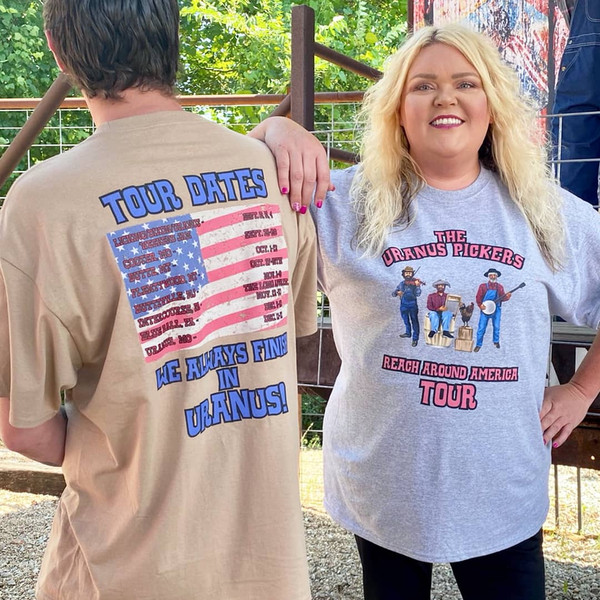 Two people modeling the "The Uranus Pickers" t-shirt. On the right, a woman wears a gray t-shirt featuring a cartoon band in overalls and the text "The Uranus Pickers Reach Around America Tour." On the left, a man shows the back of a tan t-shirt featuring an American flag overlayed with "Tour Dates" at various cities, finished with the bold blue slogan: "WE ALWAYS FINISH IN URANUS!"
