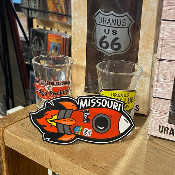 Orange rocket ship sticker displayed on a wooden shelf beside souvenir shot glasses at Uranus Fudge Factory.