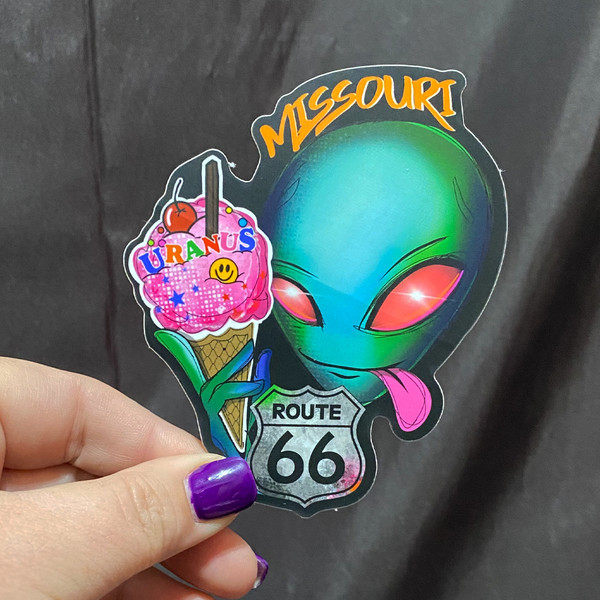 Alien ice cream sticker held by a hand with purple fingernails against a dark cloth backdrop by Uranus Fudge Factory.
