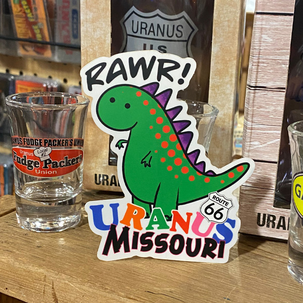 Green cartoon dinosaur sticker displayed on a wooden shelf beside a custom shot glass at Uranus Fudge Factory.