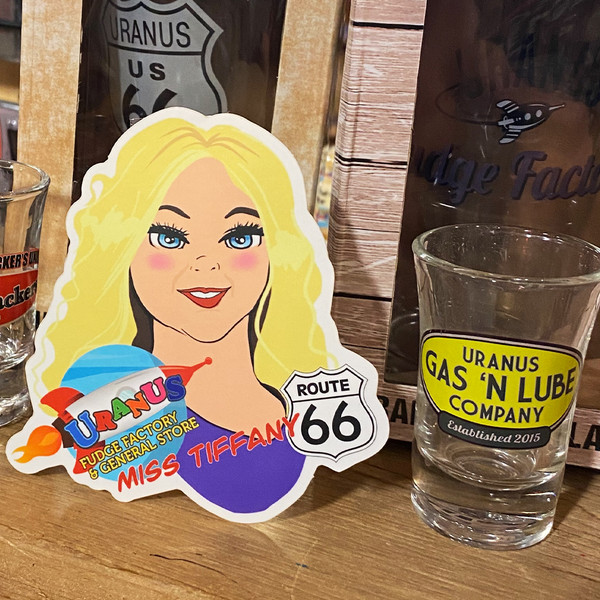 Miss. Tiffany sticker displayed on a wooden shelf beside a custom shot glass at Uranus Fudge Factory.