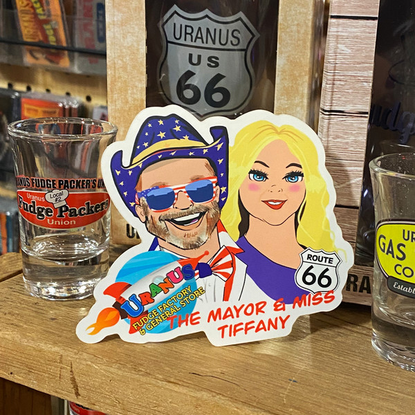 Novelty Mayor and Miss. Tiffany sticker displayed on a wooden shelf next to merchandise at Uranus Fudge Factory.