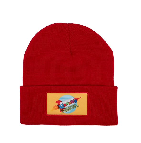 A bright red knit cuffed beanie hat against a plain white background. The front cuff features a sewn-on rectangular yellow patch. The patch displays a light blue planet with a retro red and white rocket ship flying across it, leaving a trail of orange flames.