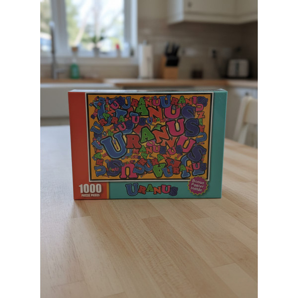 Front of the Uranus 1,000-piece jigsaw puzzle box featuring a colorful collage of the word ‘Uranus’ in overlapping fonts with a bonus poster sticker.