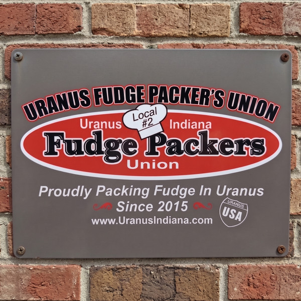 Uranus Fudge Packer's Union 12x16 metal sign mounted on a red brick wall, featuring the red and white Fudge Packers Union oval logo, Local #2 chef's hat, and "Proudly Packing Fudge In Uranus Since 2015" tagline.