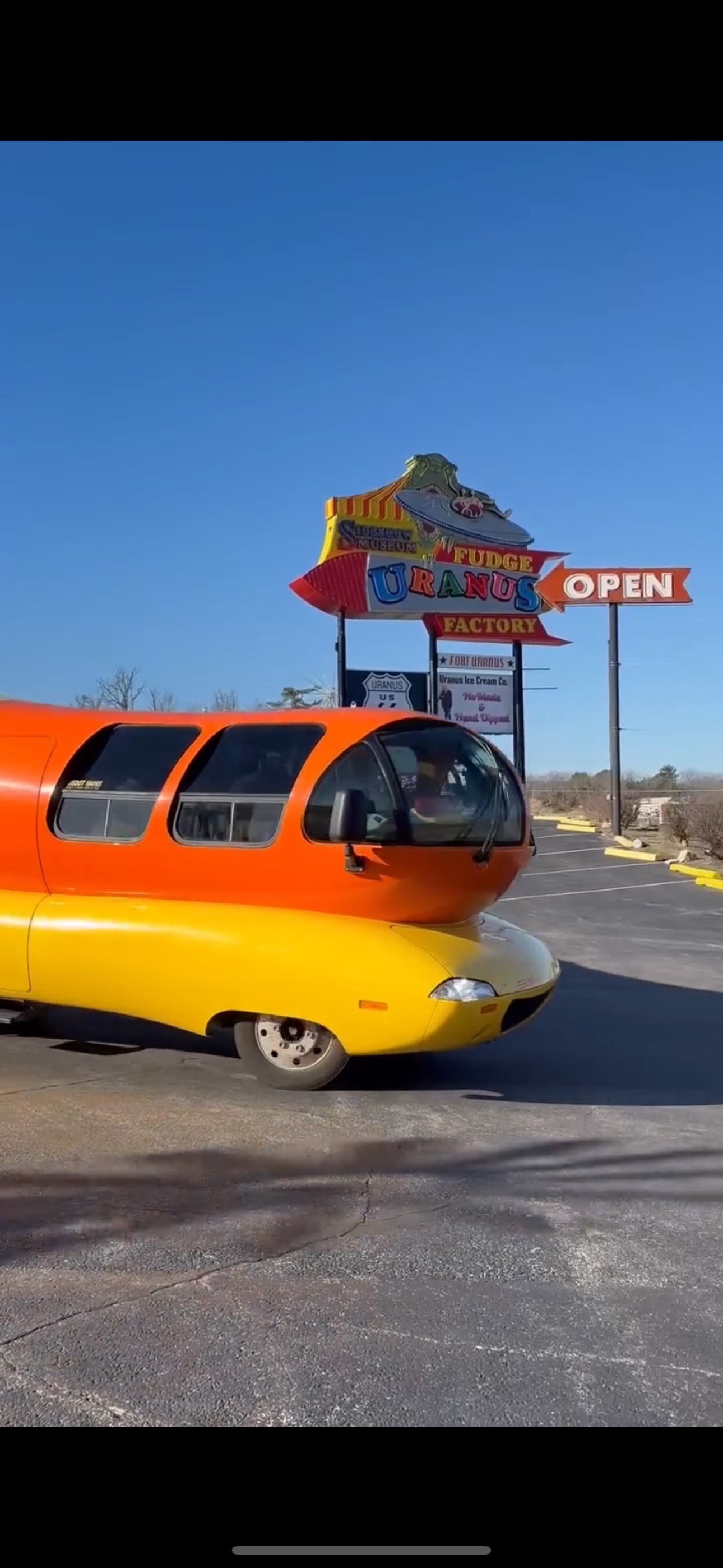 A hot dog-shaped "Wienermobile" parked in front of the Uranus Fudge Factory Missouri sign.