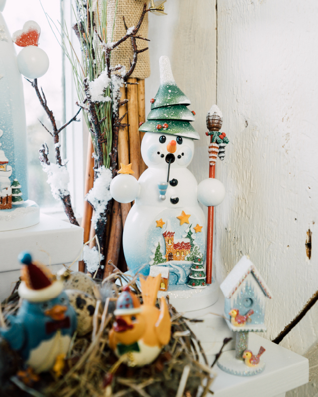 Snowman incense smoker with tree shapped hat and pinecone topped staff sitting on windowsill with a glitter frosty birdhouse ornament  and a next with two bird wood ornaments one blue and one yellow