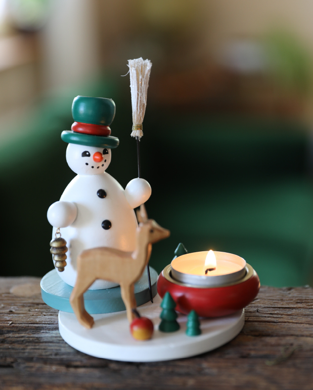 A Käthe original tealight candleholder with a deer, small trees, and an apple next to a round snowman with green and red top hat and broom in hand.