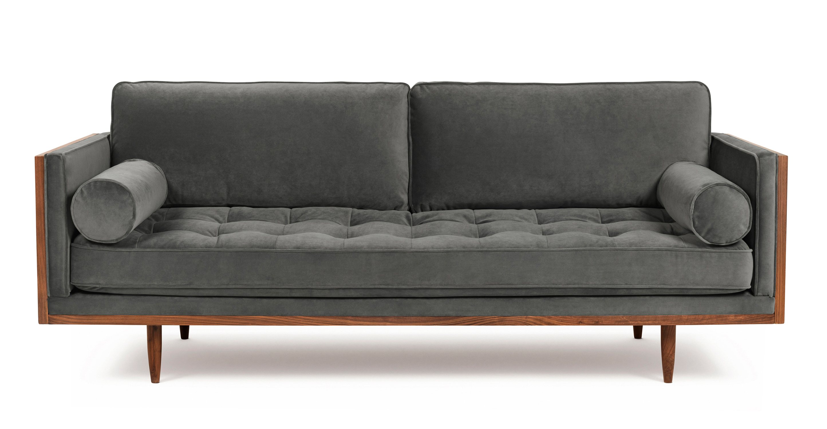 Woodrow Skandi 3 Seat Sofa in Pepper Velvet has two cylindrical bolster pillows. The sofa has one large removable seat cushion with buttonless tufting, two large removable back cushions without tufting. Sofa's frame and legs are walnut colored. The back and outsides of the sofa are wooden. The fabric color is grey.  