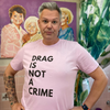 Person proudly wearing a black “Drag Is Not a Crime” shirt with bold rainbow lettering, standing in front of a graffiti wall.