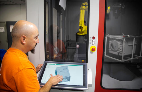 A man in an orange shirt touches a touchscreen with a 3-D model displayed.