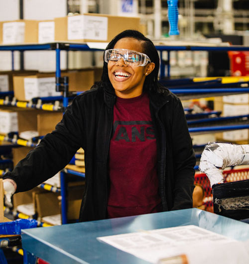 A Black woman working in a GE warehouse wears safety goggles and smiles.