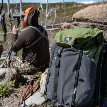 a black and avocado green Silverton backpacks leans against a boulder, nearby a man  wearing orange and camo looks through binoculars 