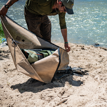 a man on a riverbank unzips the side zipper on a wetlands backpack 