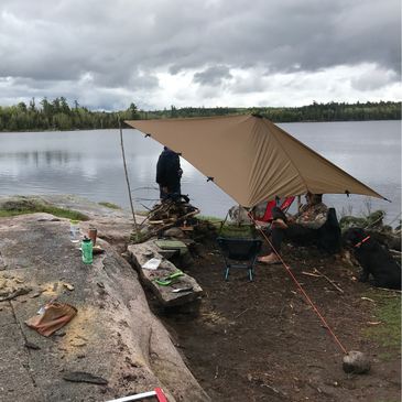 DST tarp set up in elevated diamond shape at a lakeside, a man stands under the tarp, and another man sits under the tarp in one of camp chairs