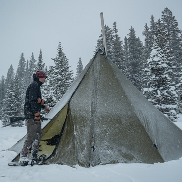 hunter wearing slow shoes enters a snow covered 12 person tipi tent with stove and gear inside